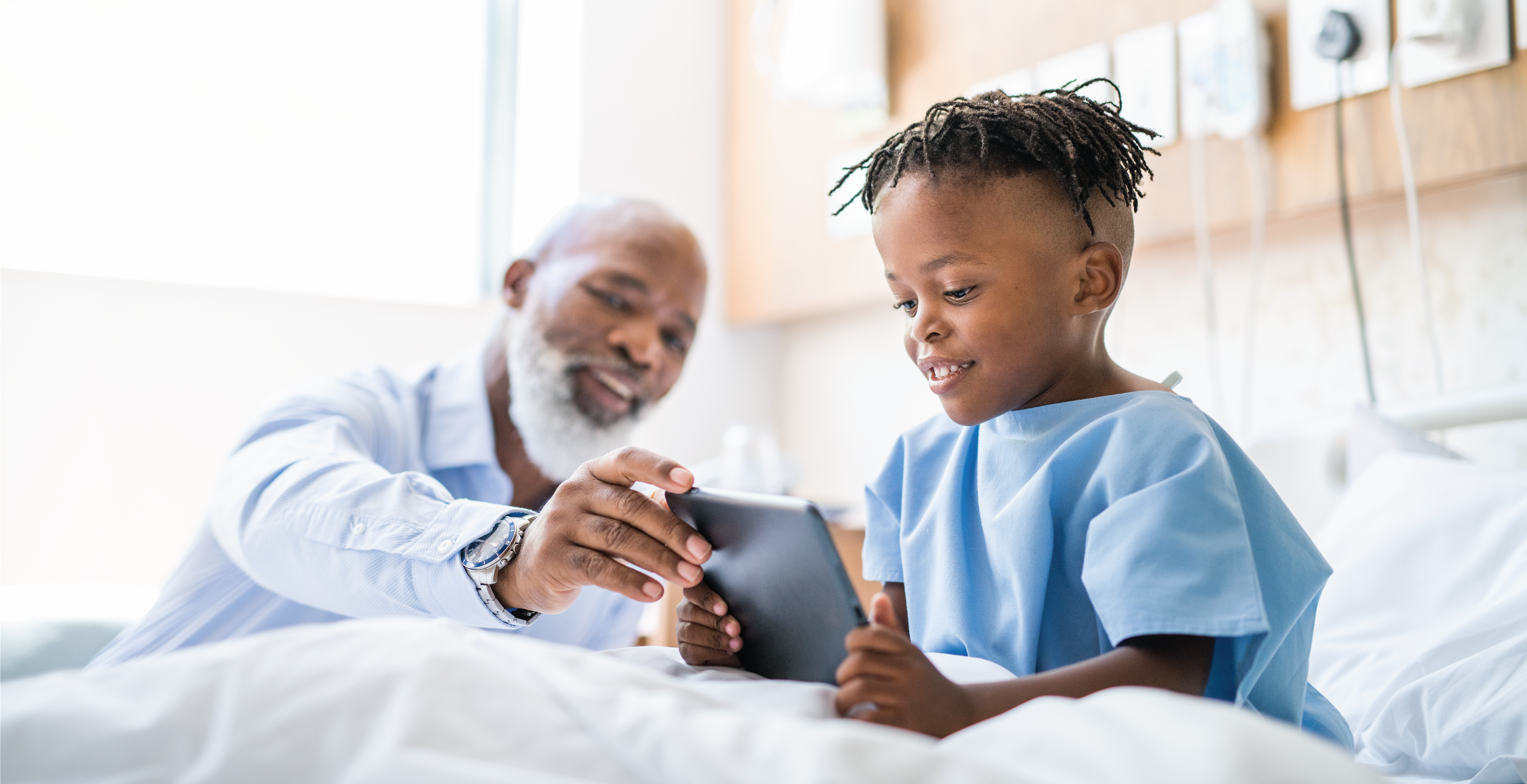 Doctor showing digital tablet to sick patient. Smiling boy patient is using wireless computer on bed. They are at hospital ward.